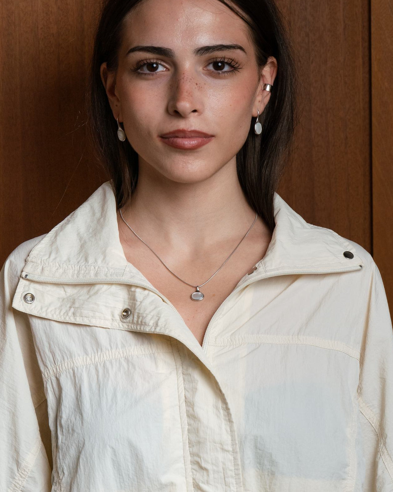 Woman wearing a light-colored jacket against a wooden background wearing Sigrid oval necklace, sideways shell cabochon on fine snake chain, rhodium or 18k gold plating.
