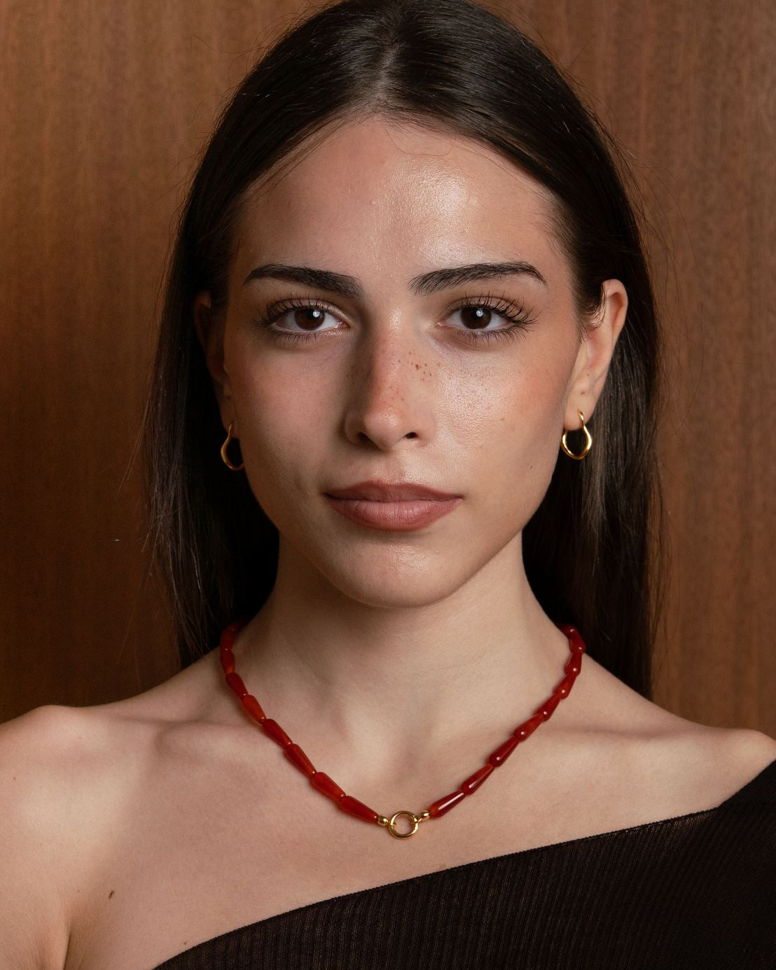Woman wearing a black off-shoulder top and red charm holding necklace and 18k gold plated flo oval hoops